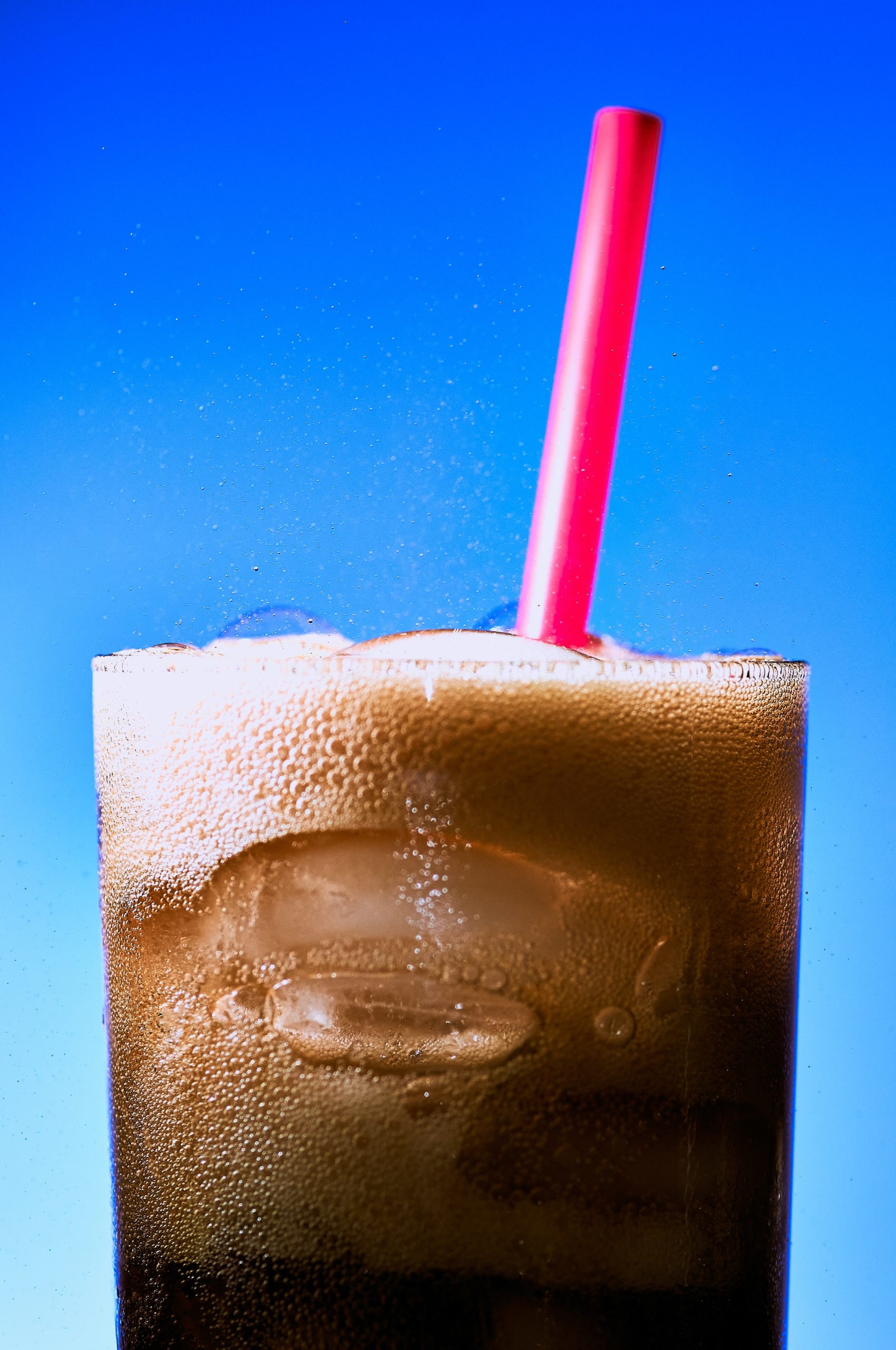 Glas voll kalter Cola mit Schaum und rotem Strohhalm, vor einem blauen Hintergrund.