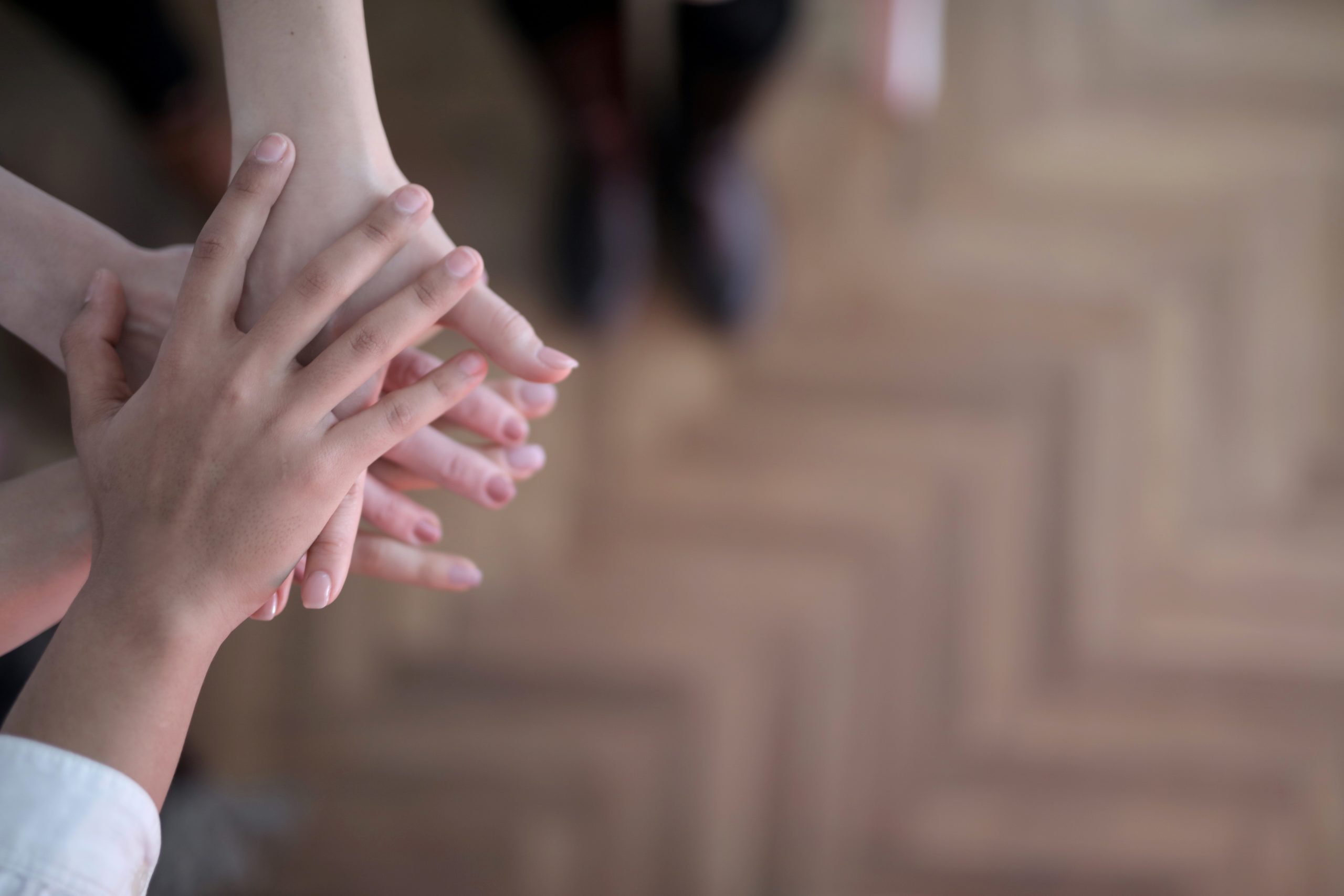 Drei Hände von Kindern sowie älteren Personen liegen übereinander, um Zusammenhalt zu symbolisieren. Im Hintergrund sind Beine in einer schwarzen Hose sowie ein dunkler Holzfußboden zu erkennen.