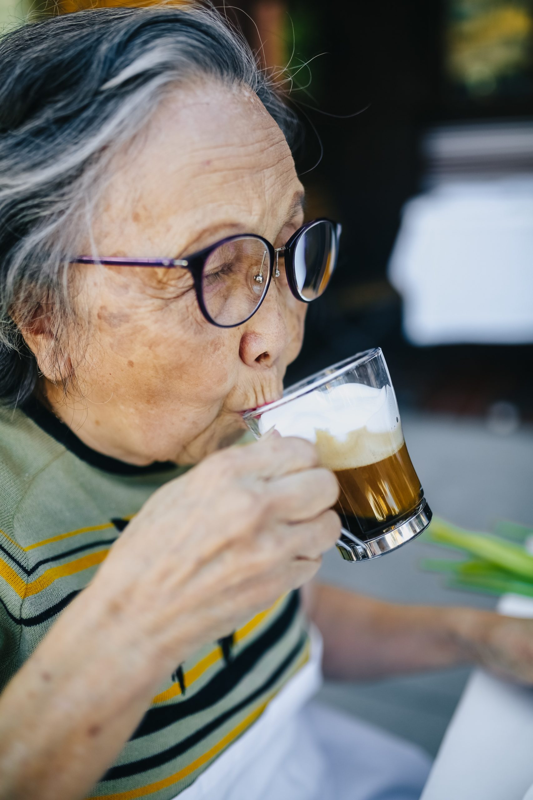 Seniorin mit großer Hornbrille trinkt Kaffee aus einem Glas.
